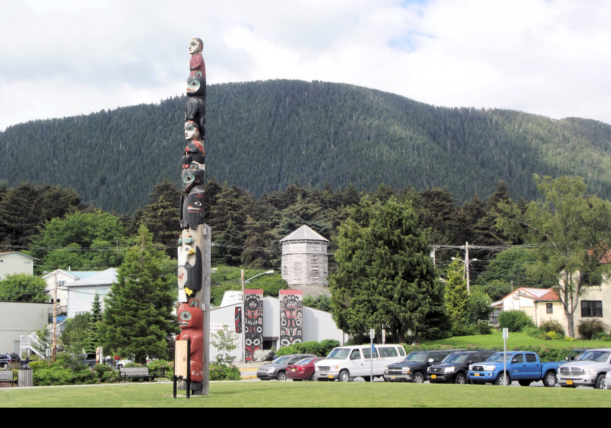 A totem pole in the city with a tribal house behind it, and a replica of one of the Russian Block Houses raised behind that.